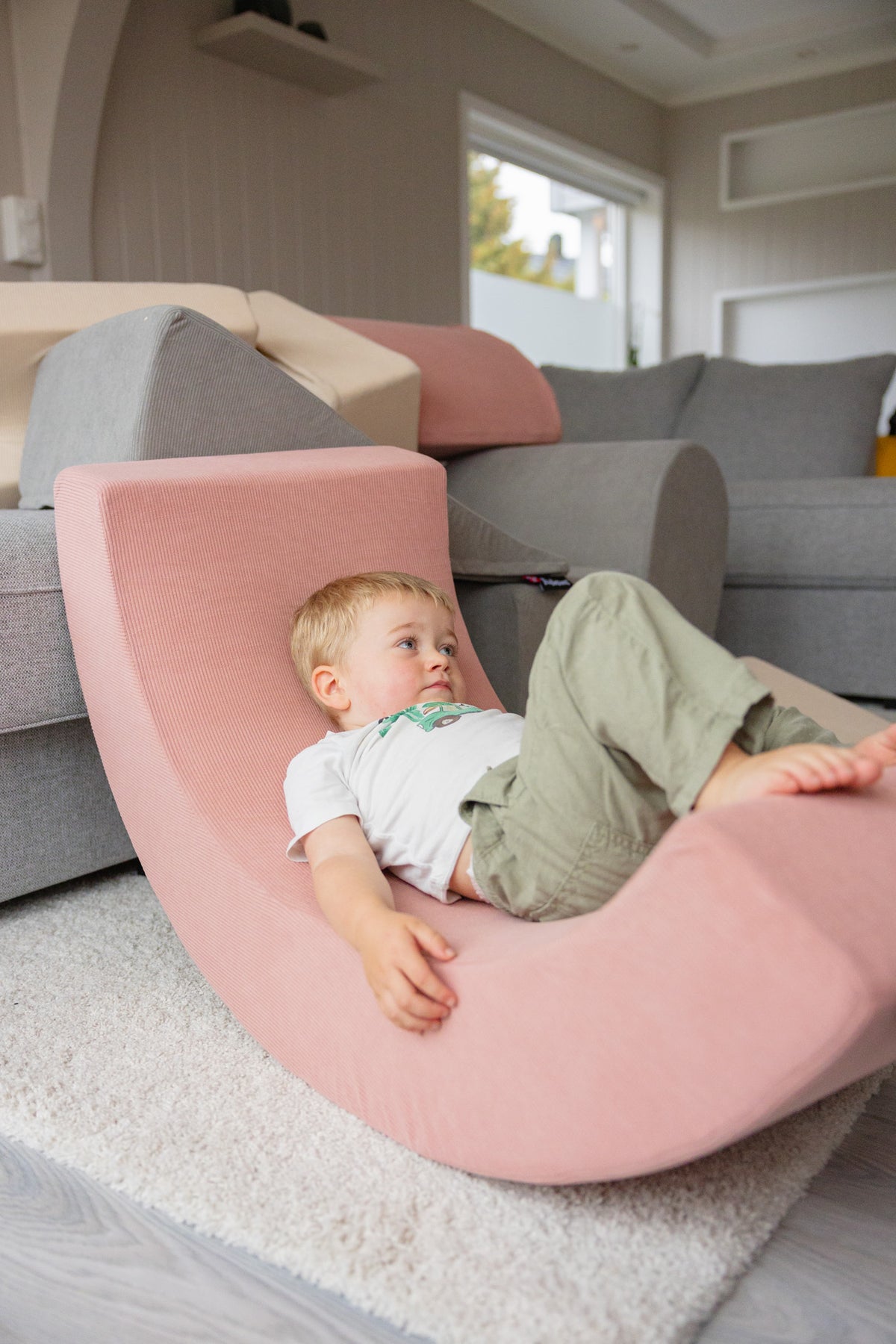 Blondt småbarn ligger avslappet i rosa Trollefant Skumregnbue i cord på hvit rye, med grå sofa og vindu i bakgrunnen