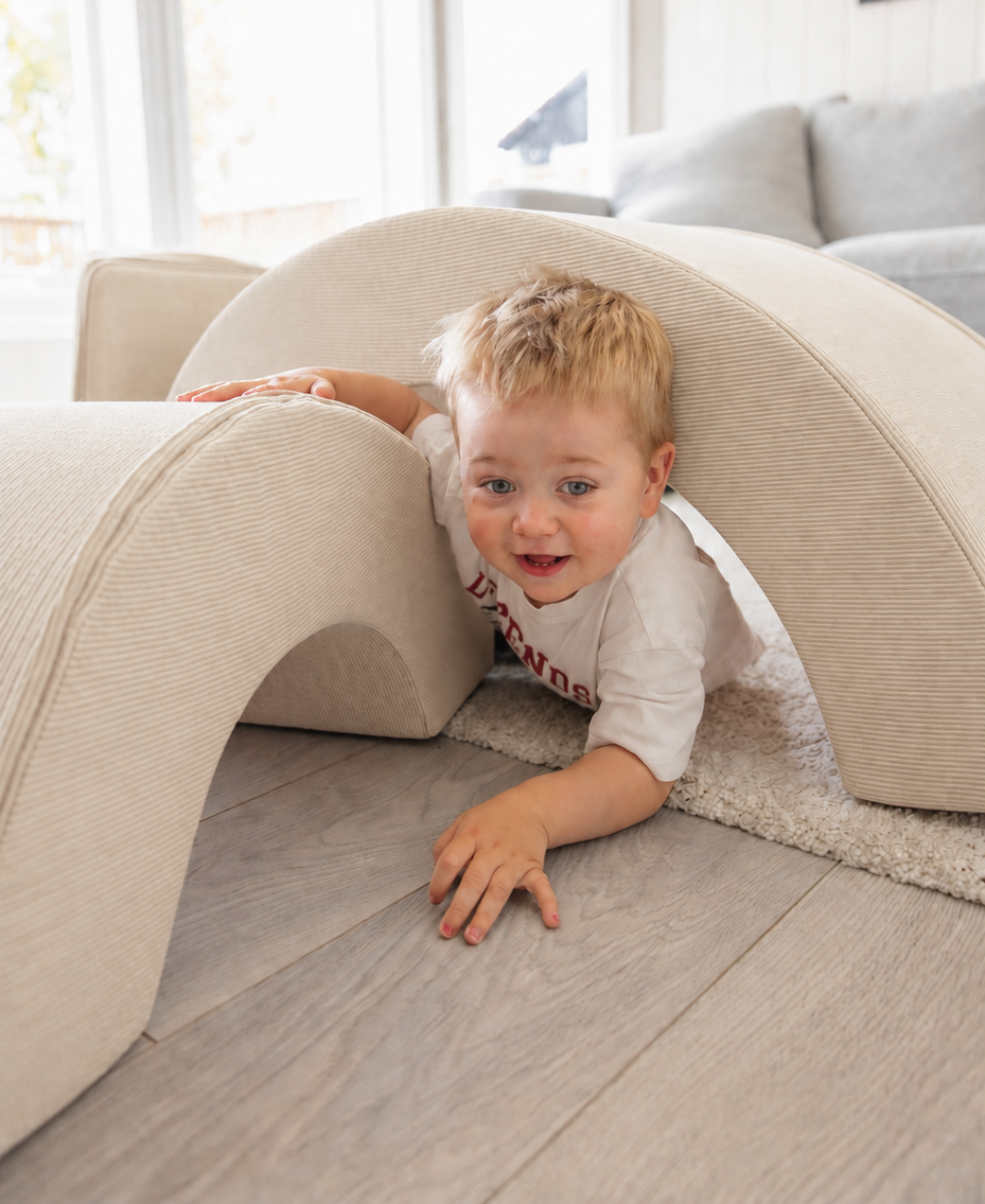Smilende blond gutt kryper gjennom buen på Trollefant Skumregnbue i beige cord på stuegulv foran sofa