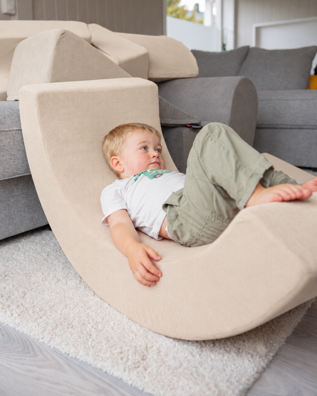 Blond gutt slapper av i Trollefant skumregnbue i beige cord på hvit rye foran grå sofa med flere skumdeler